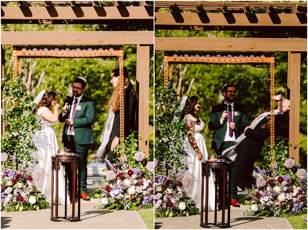 Bride in a white gown and groom in a green suit stand under a wooden arbor, the groom speaking into a microphone as a woman in black and veil looks on; colorful flowers frame the ceremony in the foreground.