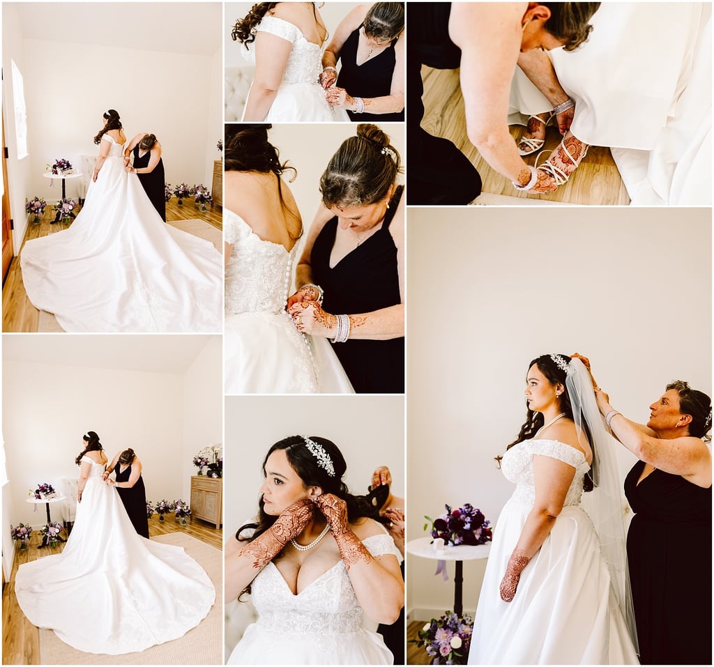 Bride in a white wedding gown standing in a bright room while a woman in black helps fasten the back of her dress; bouquets arranged on the floor nearby.