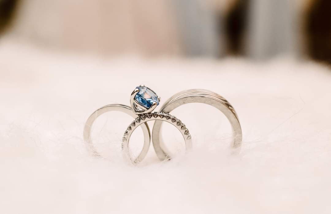 Two silver rings with a blue gemstone engagement ring resting on a soft white surface.