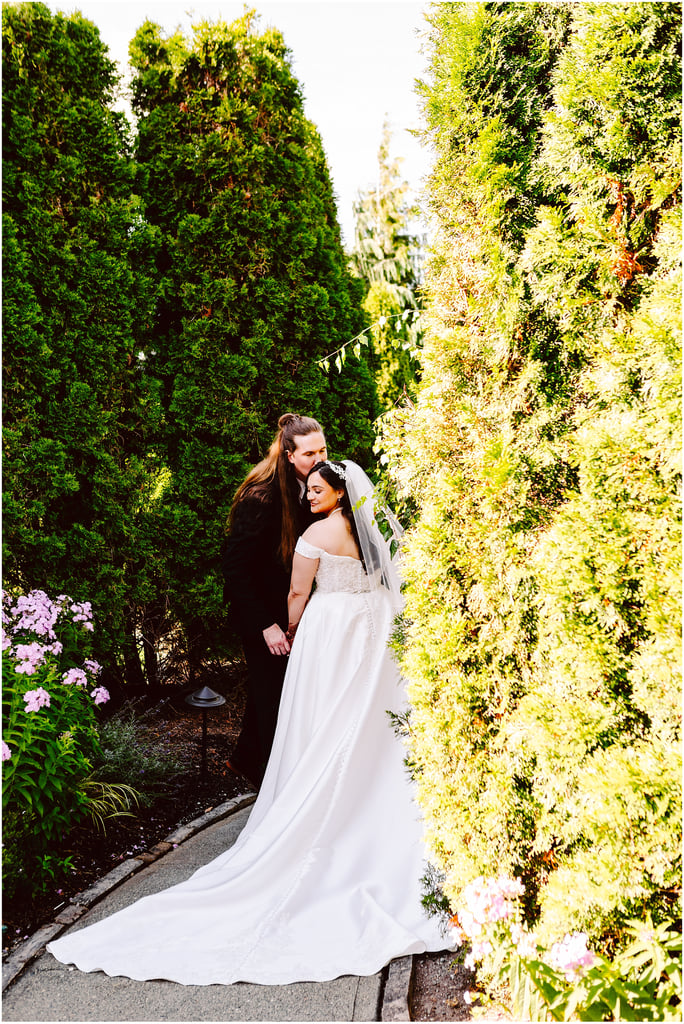 Bride in a white wedding gown and groom in a black suit kiss in a garden framed by tall green hedges and flowering plants on a sunny day