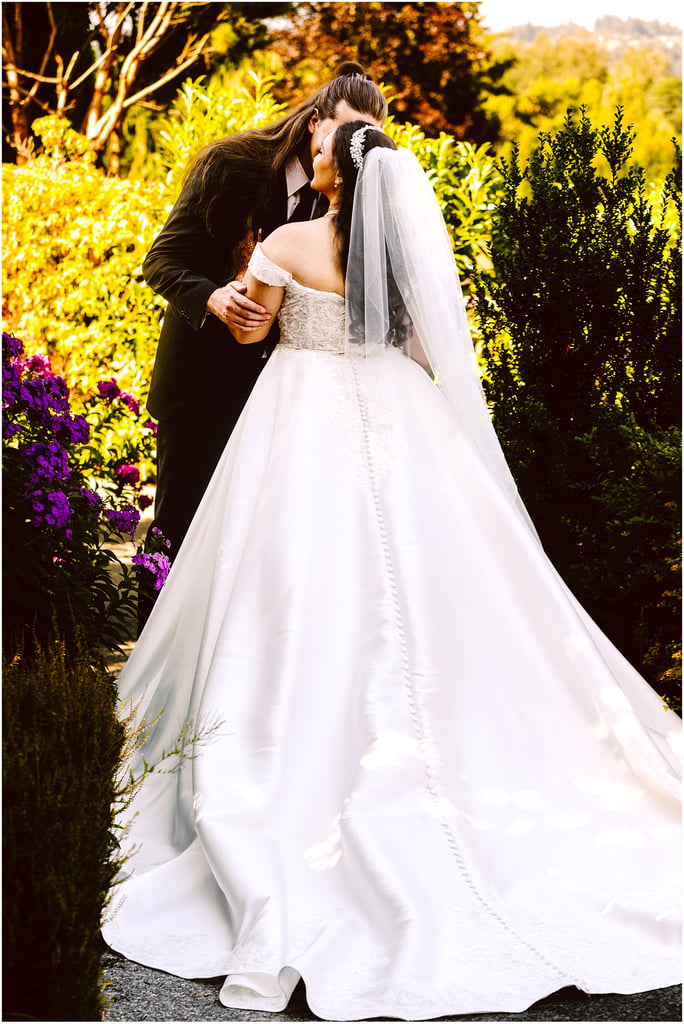 Bride and groom kissing in a sunlit garden, bride in a white wedding gown and veil among colorful flowers.