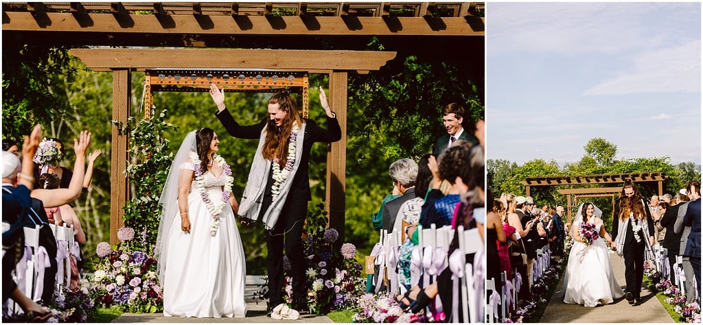 Bride in a white gown and groom in a dark suit wearing flower garlands walk under a wooden arch as guests cheer and clap. Flowers line the aisle on both sides.