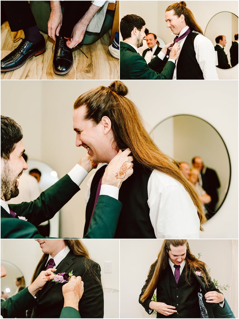 Two men in formal wear adjust a groom's purple tie, one helping while others watch in the background; henna on the helper's forearm is visible.
