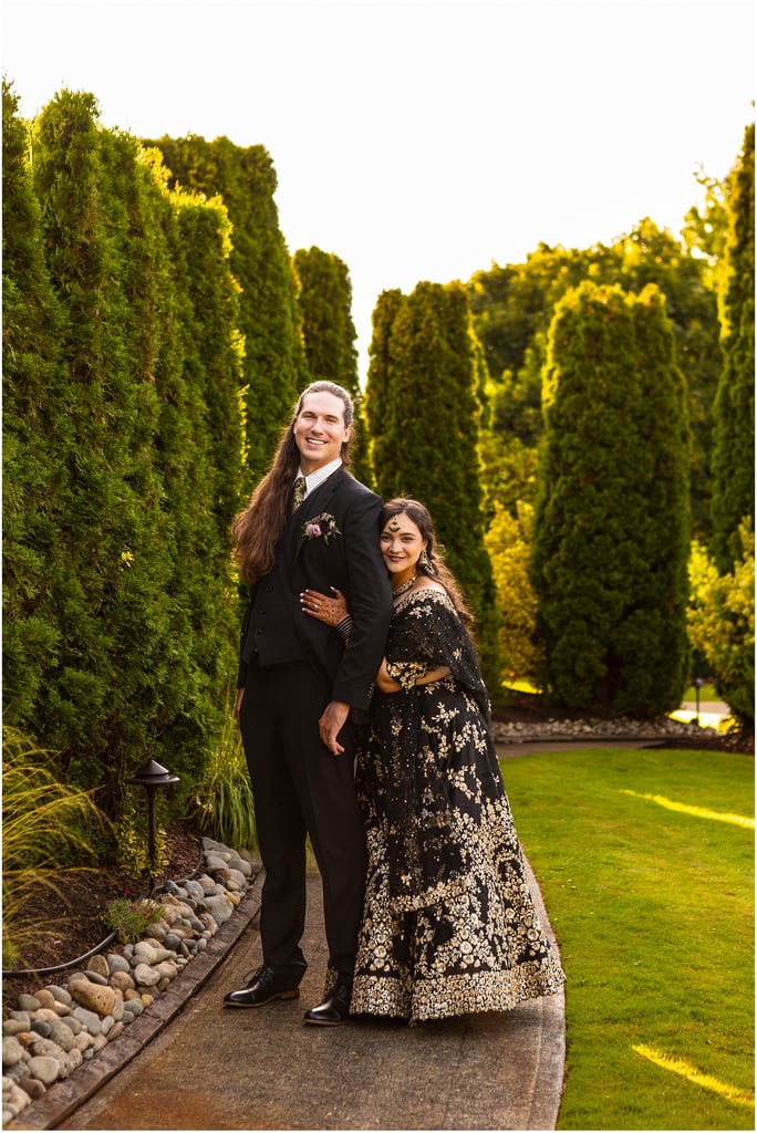 Smiling couple in formal attire standing arm-in-arm on a garden path with tall green hedges on both sides.