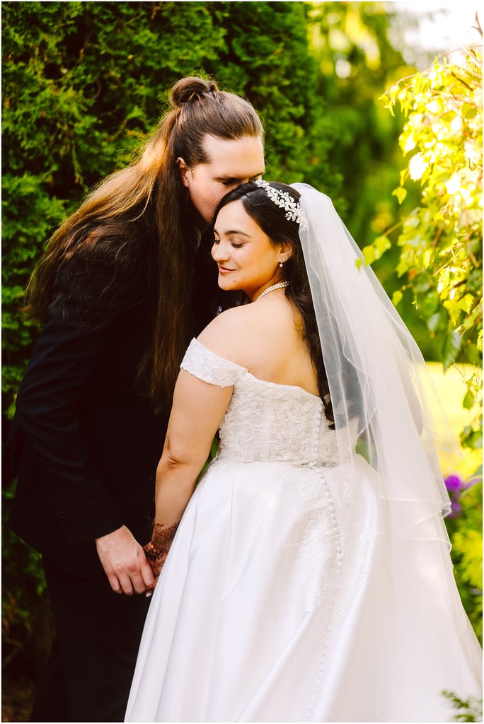 Bride in a lace wedding dress and veil stands with a long-haired groom in a garden as he kisses her head and they hold hands.