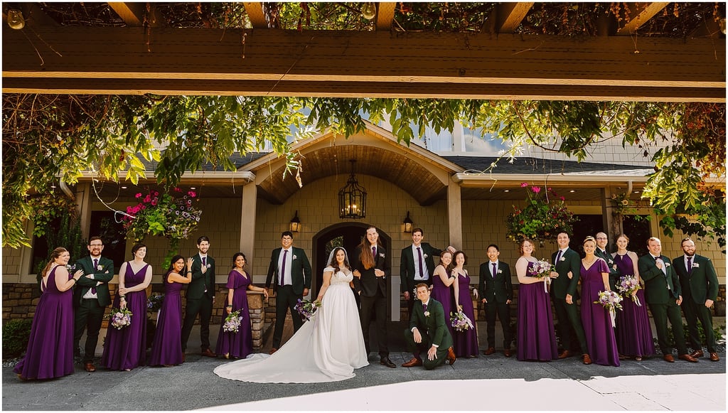 Group wedding party posed in matching purple dresses and green suits in front of a house entry.