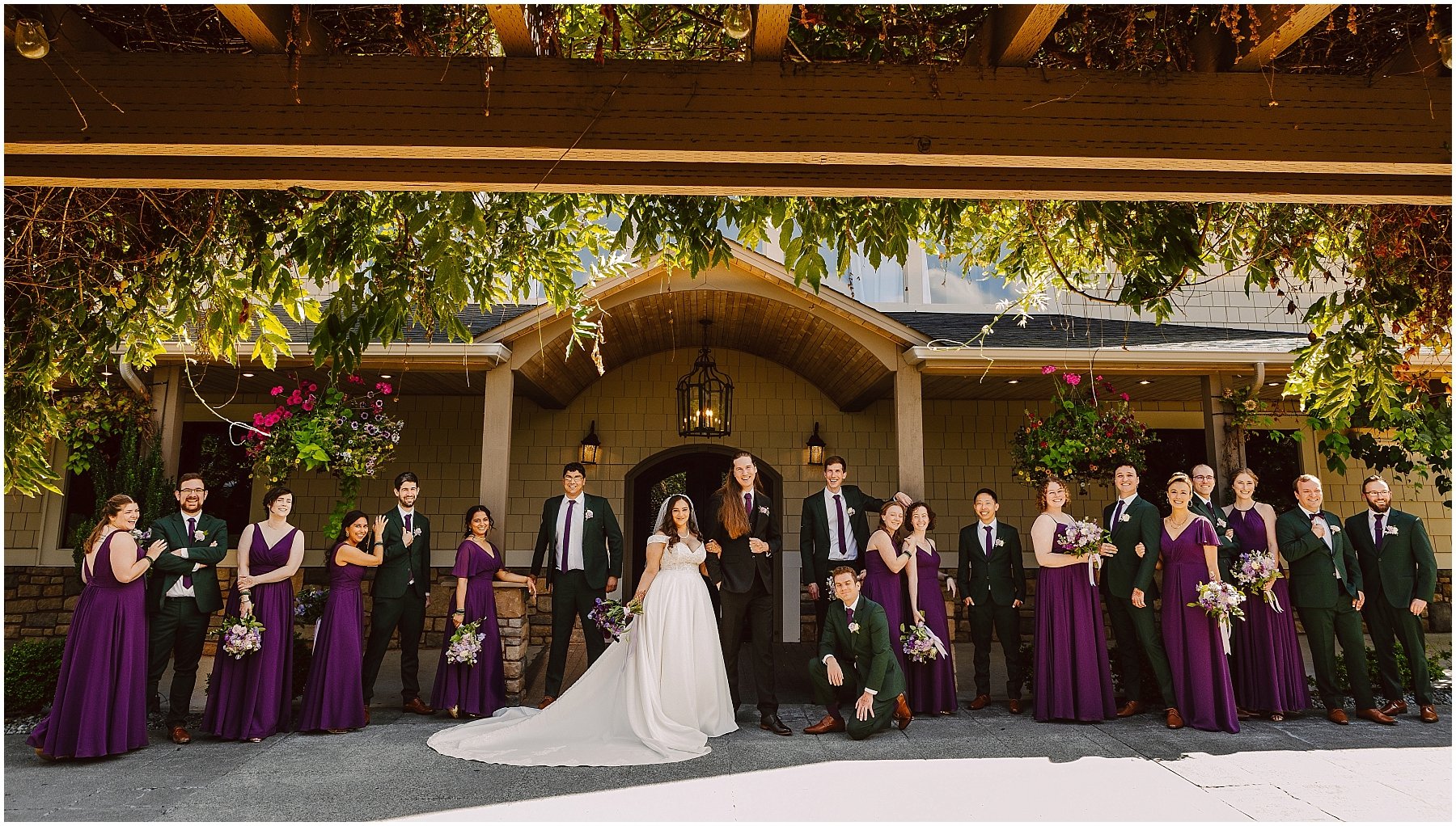 Group wedding party posed in matching purple dresses and green suits in front of a house entry.