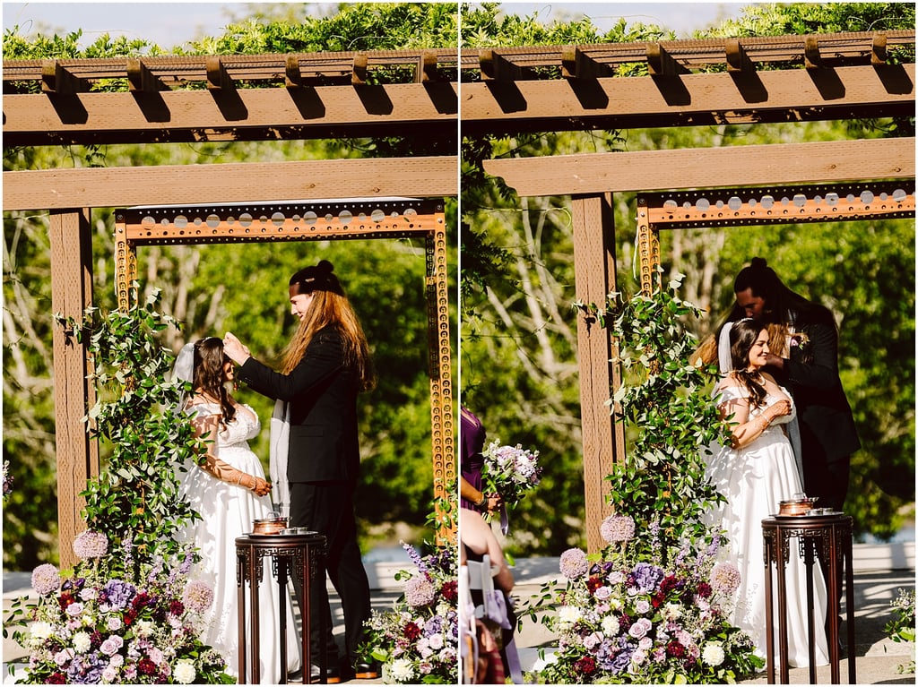 Bride in a white gown with veil stands under a wooden arbor, as a man in a black suit adjusts her hair, with lush flower arrangements nearby.