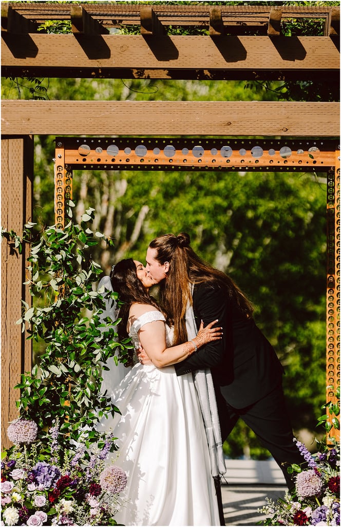 Two brides kiss beneath a rustic wooden arch framed by greenery and purple flowers at an outdoor wedding start scene.