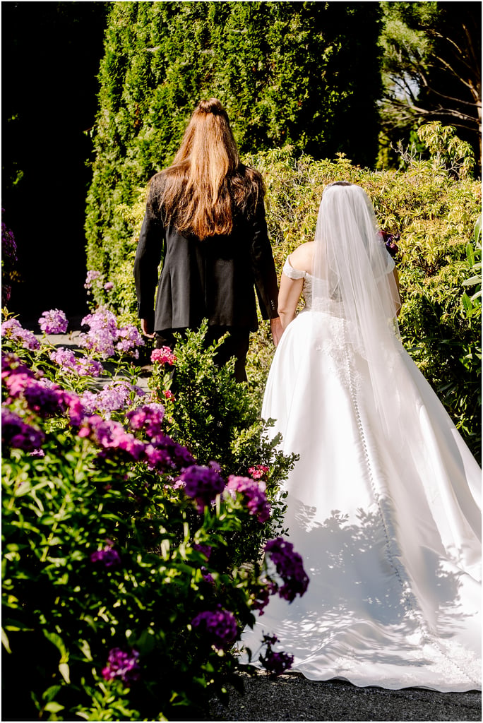 Bride in a white wedding gown and veil walks beside a man in a black suit along a garden path with vibrant purple flowers.