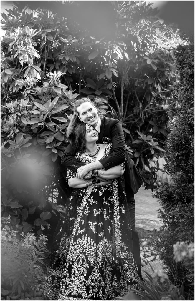 Smiling couple embracing in a garden; man in a suit behind a woman in a heavily embroidered dress, both looking happy (black and white).