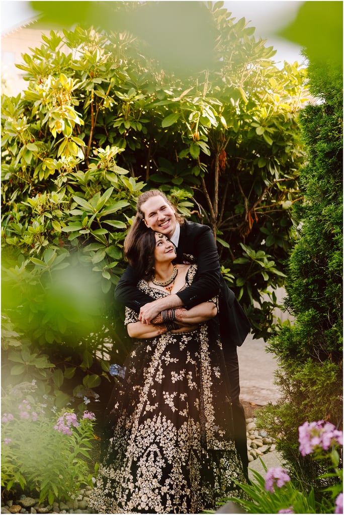 A couple poses arm-in-arm in a garden among dense green shrubs, the woman in a black embroidered dress and the man in a suit, smiling at the camera.