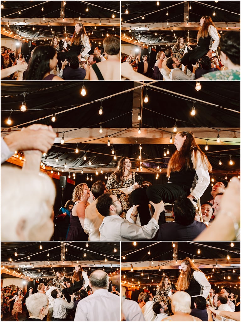Joyful wedding reception as guests hoist a person on a chair amid warm string lights under a decorated ceiling.