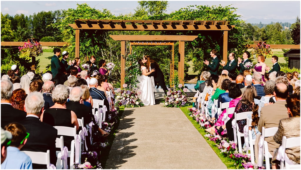 Bride and groom kiss under a wooden arbor at an outdoor wedding, surrounded by rows of seated guests and floral arrangements on the aisle sides.
