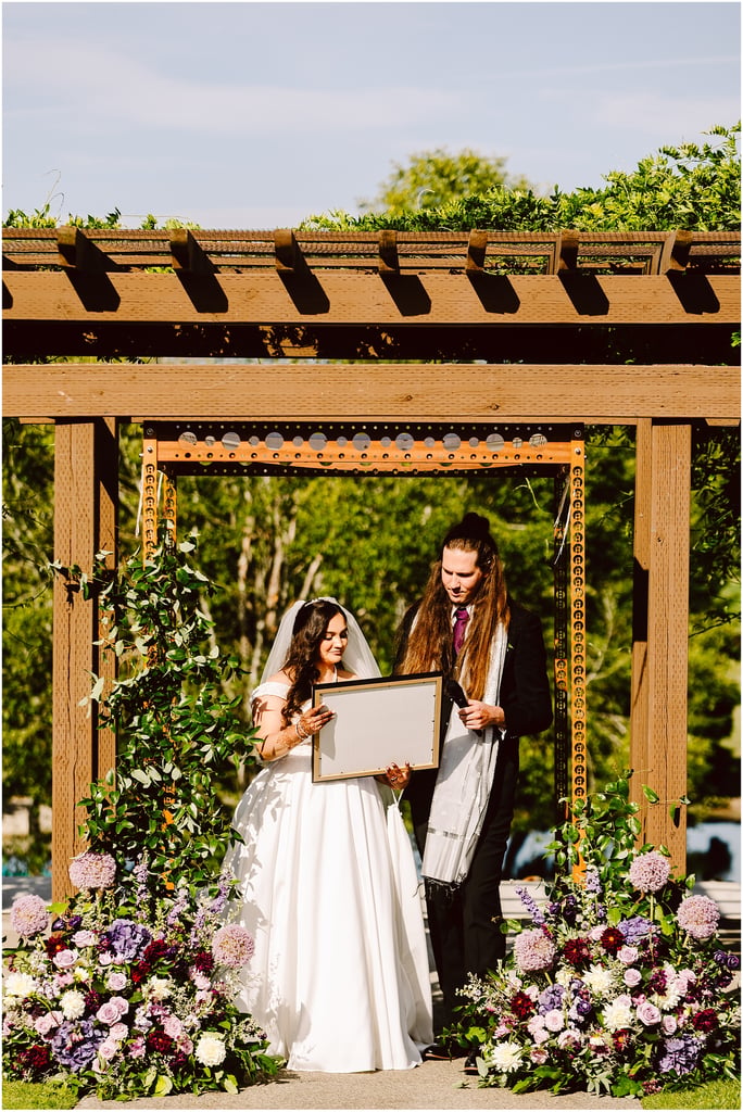 Bride and groom under a wooden pergola, the bride holding a framed board amid vibrant pink, purple, and white flowers.