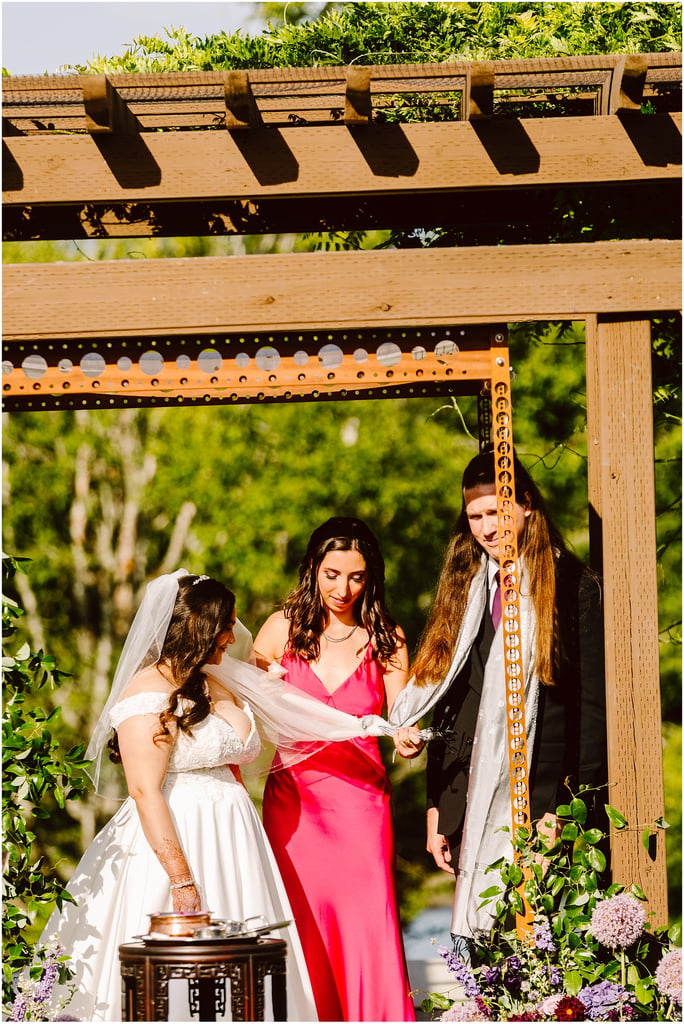 Bride in a white wedding dress with veil standing under a wooden arbor while a woman in a pink dress helps adjust her veil, outdoors with flowers nearby.