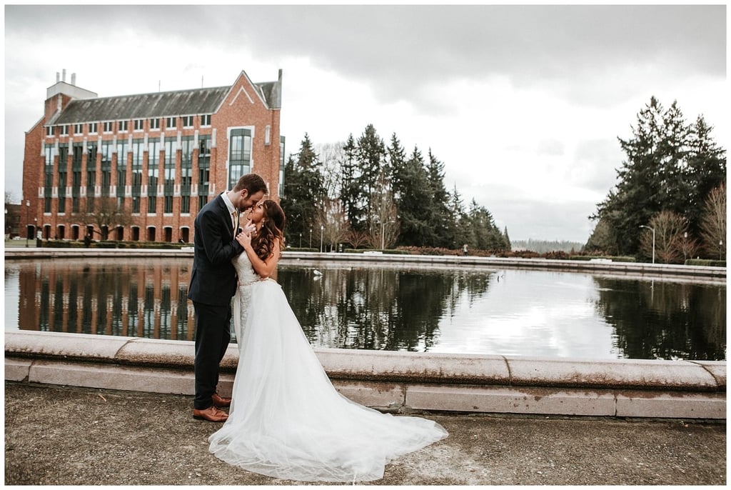 Premier Snohomish & Seattle Wedding Photographer | GSquared Weddings Photography A bride and groom stand close together, embracing beside a reflective pond at a Snohomish Seattle wedding, with a large brick building and trees in the background under a cloudy sky. 47.9129° N, 122.0982° W | Serving Snohomish, Seattle , Orlando & Beyond