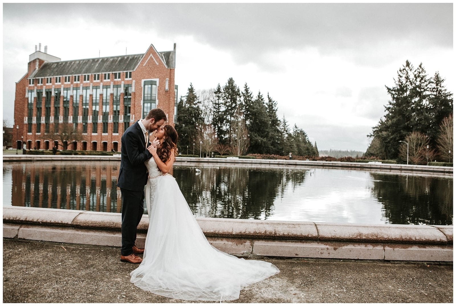 Premier Snohomish & Seattle Wedding Photographer | GSquared Weddings Photography A bride and groom stand close together, embracing beside a reflective pond at a Snohomish Seattle wedding, with a large brick building and trees in the background under a cloudy sky. 47.9129° N, 122.0982° W | Serving Snohomish, Seattle , Orlando & Beyond