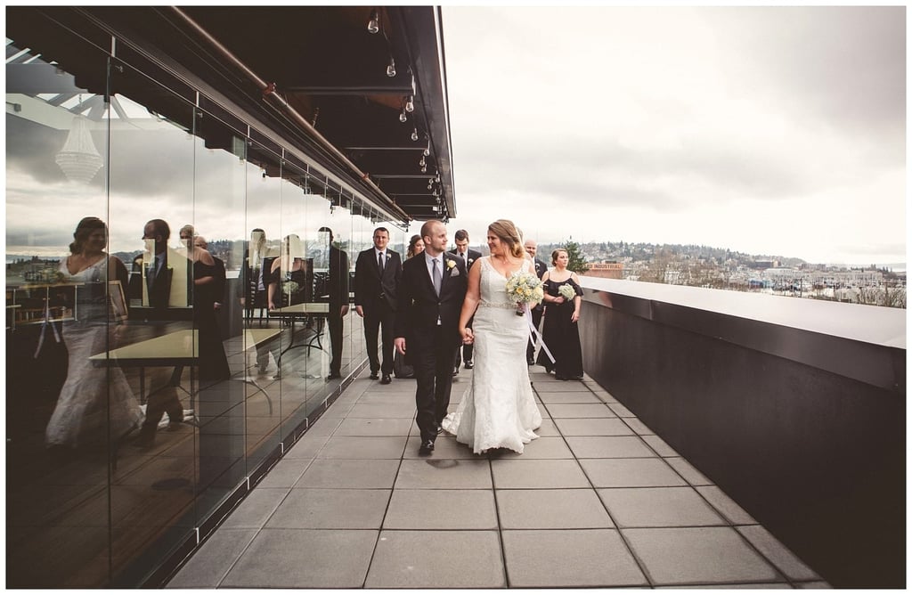 Premier Snohomish & Seattle Wedding Photographer | GSquared Weddings Photography A bride and groom walk together on a rooftop terrace in Snohomish, smiling and holding hands, while bridesmaids and groomsmen follow behind. The scene is beautifully reflected in the glass wall beside them under a cloudy Seattle sky. 47.9129° N, 122.0982° W | Serving Snohomish, Seattle , Orlando & Beyond