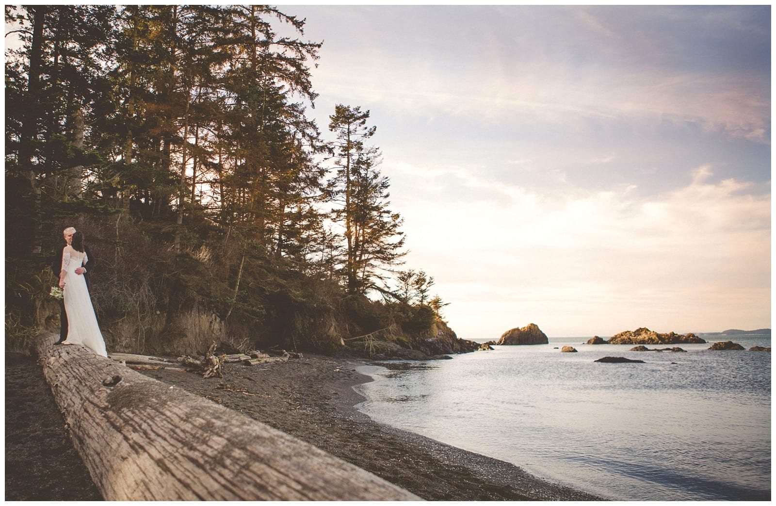 Premier Snohomish & Seattle Wedding Photographer | GSquared Weddings Photography A bride in a white dress stands on a quiet Snohomish beach near tall trees, holding a bouquet. The sky is soft and pastel as the sun sets over the calm ocean and distant rocks—perfect for a Seattle wedding engagement. 47.9129° N, 122.0982° W | Serving Snohomish, Seattle , Orlando & Beyond