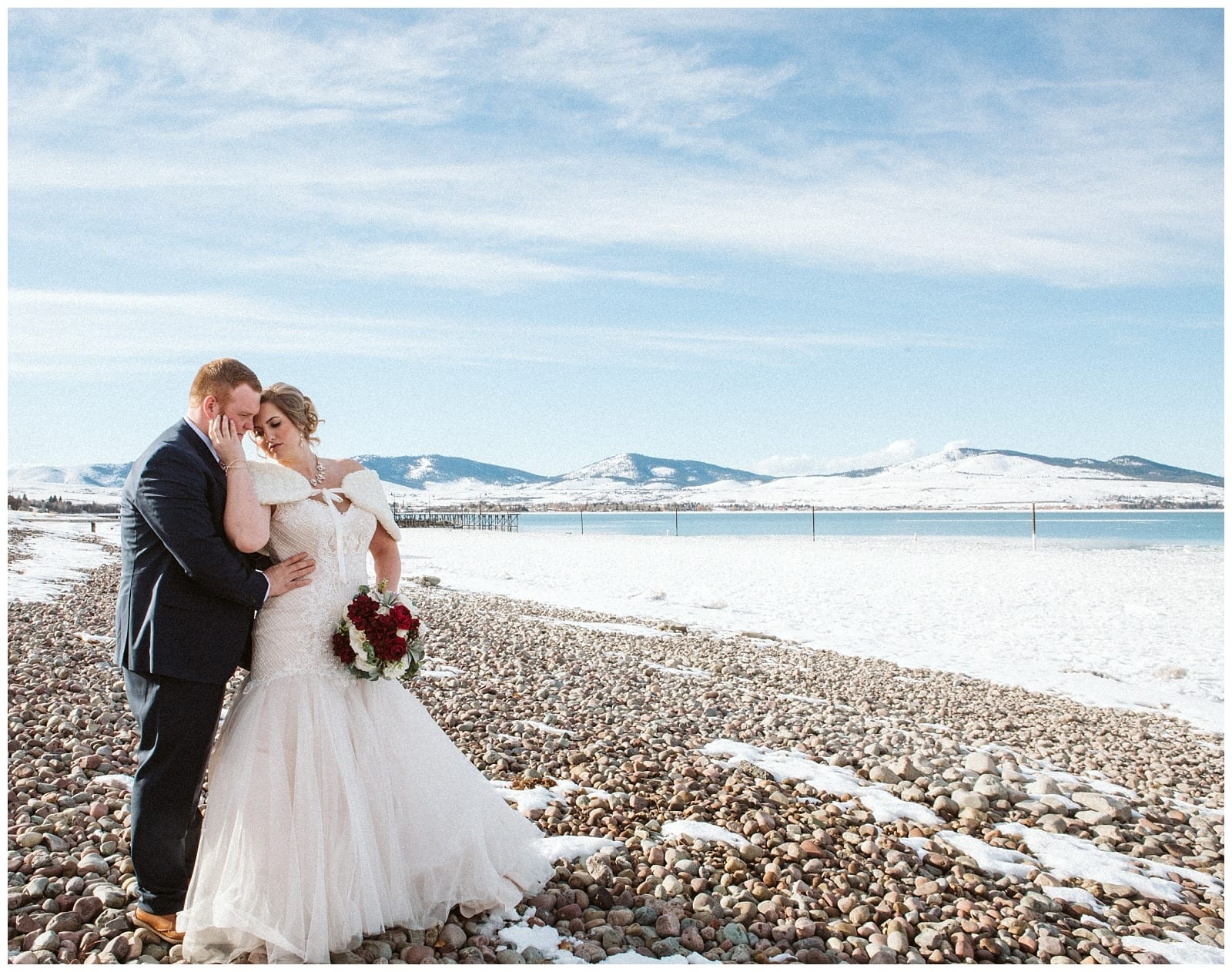 Premier Snohomish & Seattle Wedding Photographer | GSquared Weddings Photography A bride and groom embrace on a snowy, rocky beach with a lake and snow-covered mountains in the background under a clear blue sky, capturing the magic of a Snohomish Seattle wedding. The bride holds a bouquet of red and white flowers. 47.9129° N, 122.0982° W | Serving Snohomish, Seattle , Orlando & Beyond
