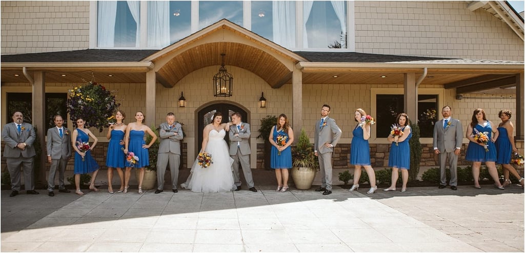 Premier Snohomish & Seattle Wedding Photographer | GSquared Weddings Photography A Snohomish wedding party poses outside a tan building. The bride and groom stand in the center, surrounded by bridesmaids in blue dresses and groomsmen in gray suits, all holding colorful bouquets and smiling. 47.9129° N, 122.0982° W | Serving Snohomish, Seattle , Orlando & Beyond