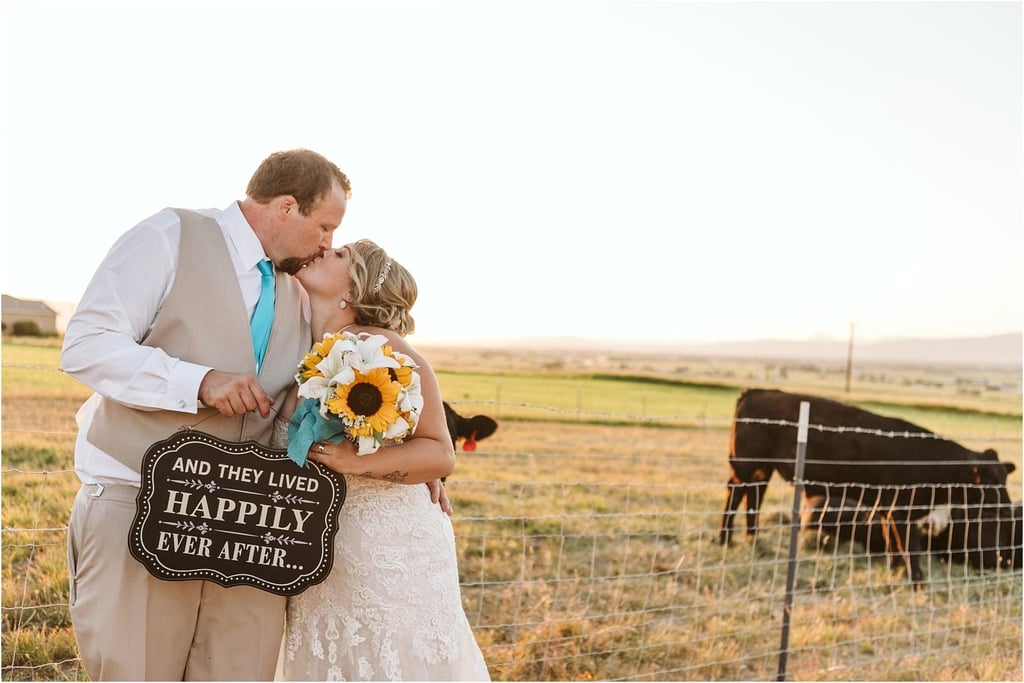 Premier Snohomish & Seattle Wedding Photographer | GSquared Weddings Photography A bride and groom kiss in a sunny Snohomish field by a wire fence, holding sunflowers and a sign reading And they lived happily ever after. Cows graze in the background—a perfect Seattle wedding engagement moment. 47.9129° N, 122.0982° W | Serving Snohomish, Seattle , Orlando & Beyond