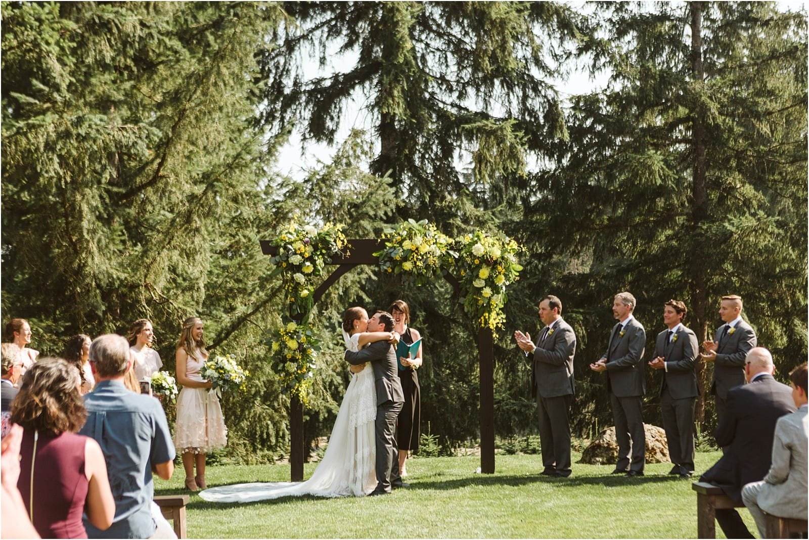 Premier Snohomish & Seattle Wedding Photographer | GSquared Weddings Photography A bride and groom embrace under a floral arch during a Snohomish outdoor wedding ceremony surrounded by trees, while their wedding party and guests watch and applaud. 47.9129° N, 122.0982° W | Serving Snohomish, Seattle , Orlando & Beyond