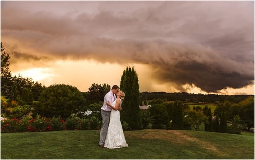 A bride and groom embrace on a grassy Snohomish hill at sunset with dramatic dark storm clouds and golden light behind them surrounded by lush trees and a scenic landscape Snohomish Wedding Photography A bride and groom embrace on a grassy Snohomish hill at sunset with dramatic dark storm clouds and golden light behind them surrounded by lush trees and a scenic landscape Snohomish Wedding Photography