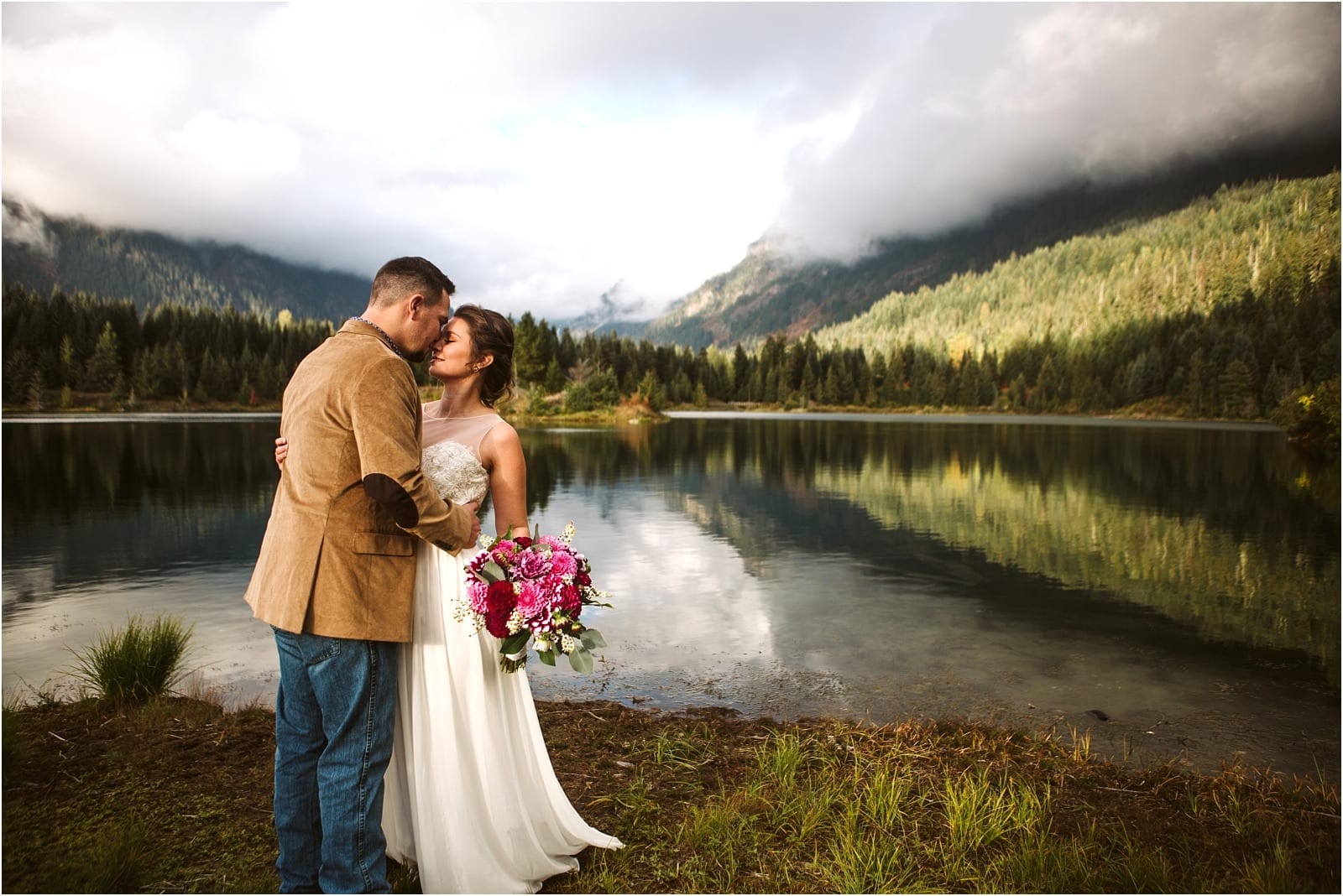 Premier Snohomish & Seattle Wedding Photographer | GSquared Weddings Photography A bride and groom stand close together by a calm lake near Snohomish, surrounded by trees and mountains, with the bride holding a bouquet of pink flowers. The partly cloudy sky creates a serene, romantic Seattle wedding scene. 47.9129° N, 122.0982° W | Serving Snohomish, Seattle , Orlando & Beyond