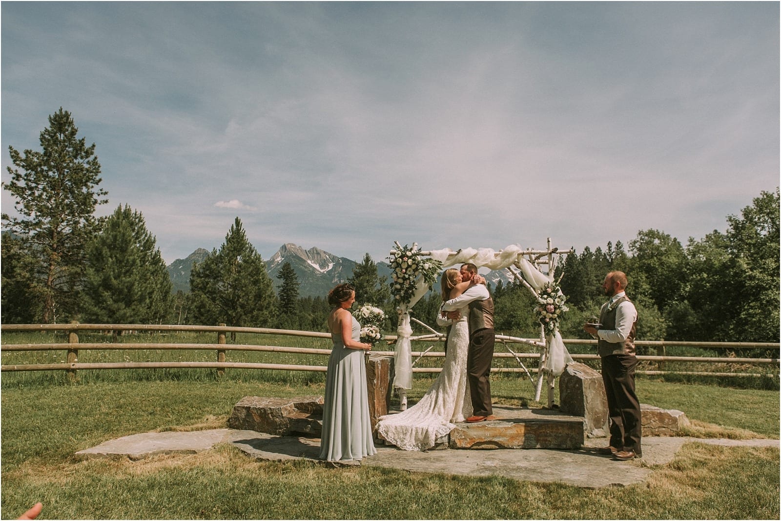 Premier Snohomish & Seattle Wedding Photographer | GSquared Weddings Photography A bride and groom share a kiss at their Snohomish outdoor wedding ceremony, standing under a decorated arch with mountains and pine trees in the background. Two attendants, one in a silver dress and one in a vest, stand nearby. 47.9129° N, 122.0982° W | Serving Snohomish, Seattle , Orlando & Beyond