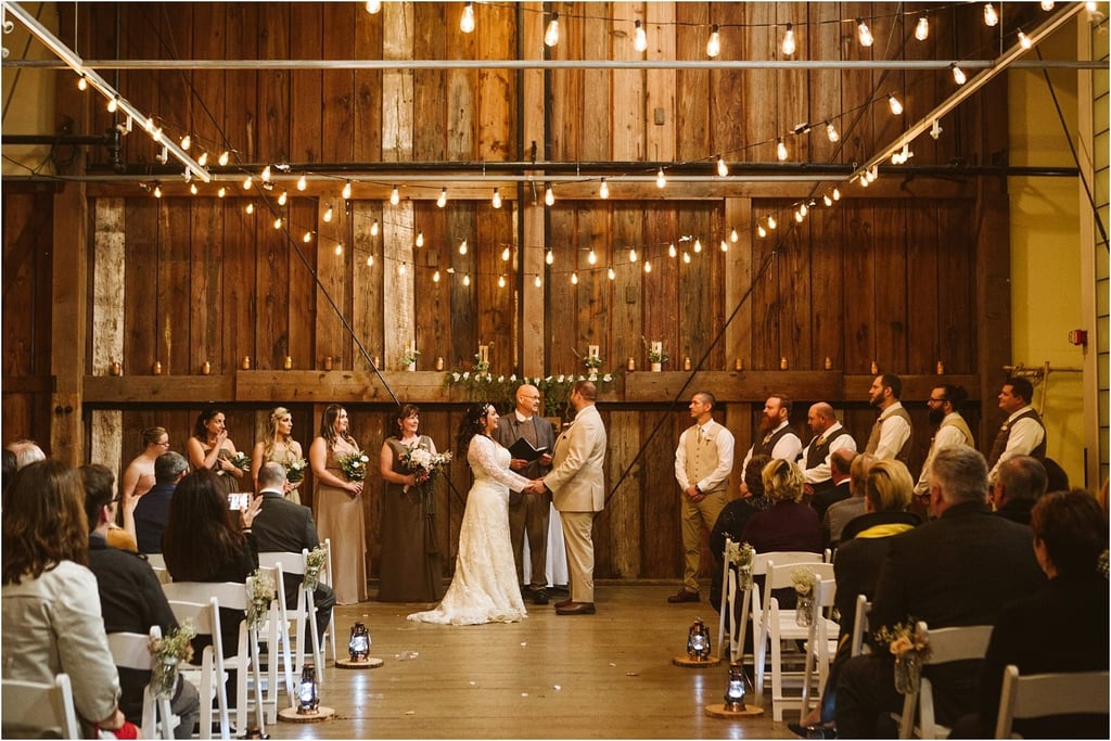 Premier Snohomish & Seattle Wedding Photographer | GSquared Weddings Photography A bride and groom stand holding hands at the altar during an indoor rustic Snohomish wedding ceremony, surrounded by their wedding party and guests, beneath string lights and in front of a wooden backdrop. 47.9129° N, 122.0982° W | Serving Snohomish, Seattle , Orlando & Beyond
