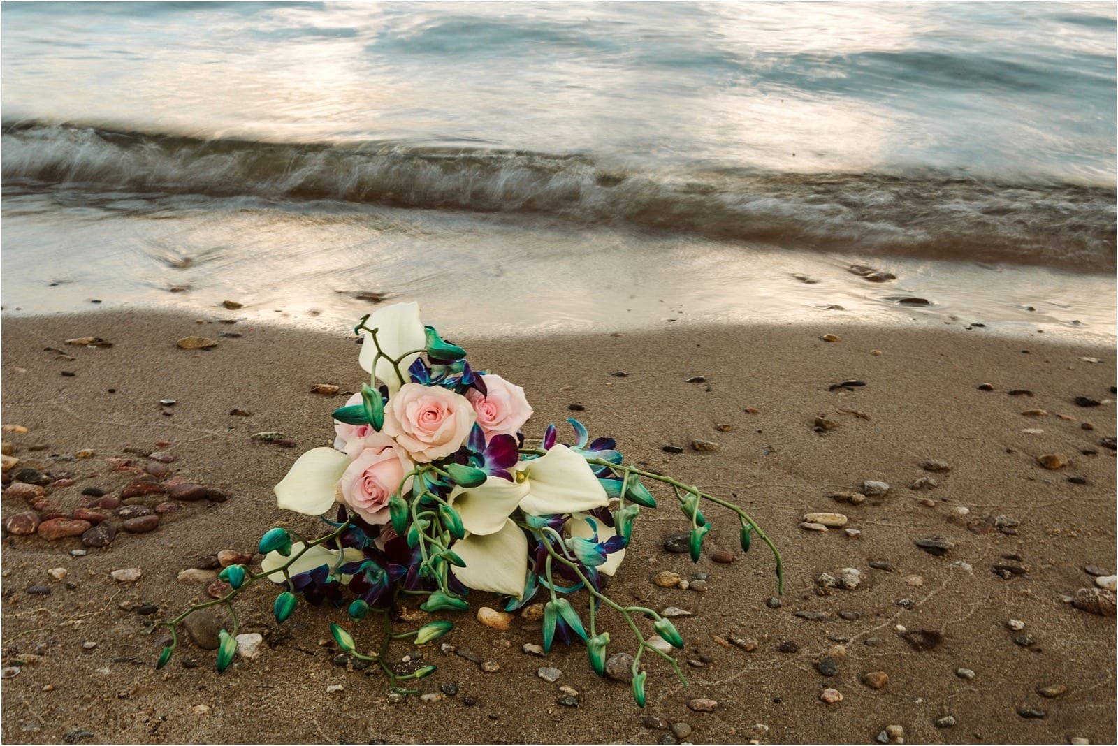 Premier Snohomish & Seattle Wedding Photographer | GSquared Weddings Photography A bouquet with pink roses, white calla lilies, and blue-purple flowers lies on a sandy beach near gentle waves—perfect for a Snohomish or Seattle wedding engagement, surrounded by small pebbles. 47.9129° N, 122.0982° W | Serving Snohomish, Seattle , Orlando & Beyond