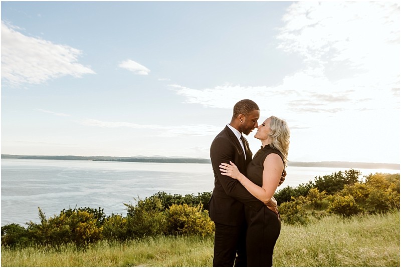 Premier Snohomish & Seattle Wedding Photographer | GSquared Weddings Photography A couple dressed in formal black attire stands closely together on a grassy hill overlooking a large body of water under a partly cloudy sky, embracing and gazing into each other's eyes during their Snohomish wedding engagement. 47.9129° N, 122.0982° W | Serving Snohomish, Seattle , Orlando & Beyond
