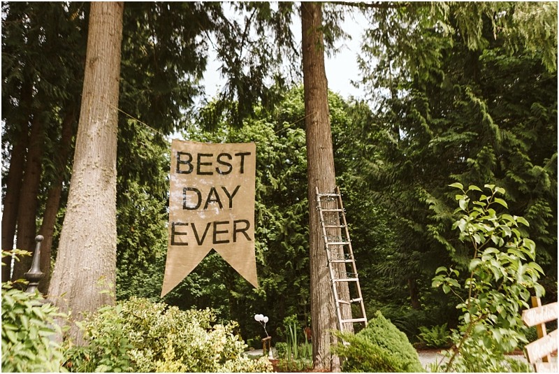 Premier Snohomish & Seattle Wedding Photographer | GSquared Weddings Photography A large burlap banner reading BEST DAY EVER hangs between two tall trees in a lush, green forested area—an idyllic scene perfect for a Snohomish Seattle wedding or engagement. A wooden ladder leans against one of the trees nearby. 47.9129° N, 122.0982° W | Serving Snohomish, Seattle , Orlando & Beyond