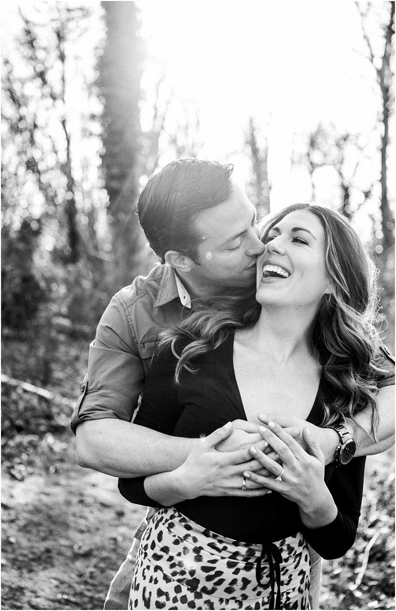 Premier Snohomish & Seattle Wedding Photographer | GSquared Weddings Photography A man embraces a smiling woman from behind, playfully kissing her cheek as they stand on a sunlit forest path. Both appear happy and relaxed, with trees and sunlight in the background—capturing a joyful Snohomish engagement in black and white. 47.9129° N, 122.0982° W | Serving Snohomish, Seattle , Orlando & Beyond