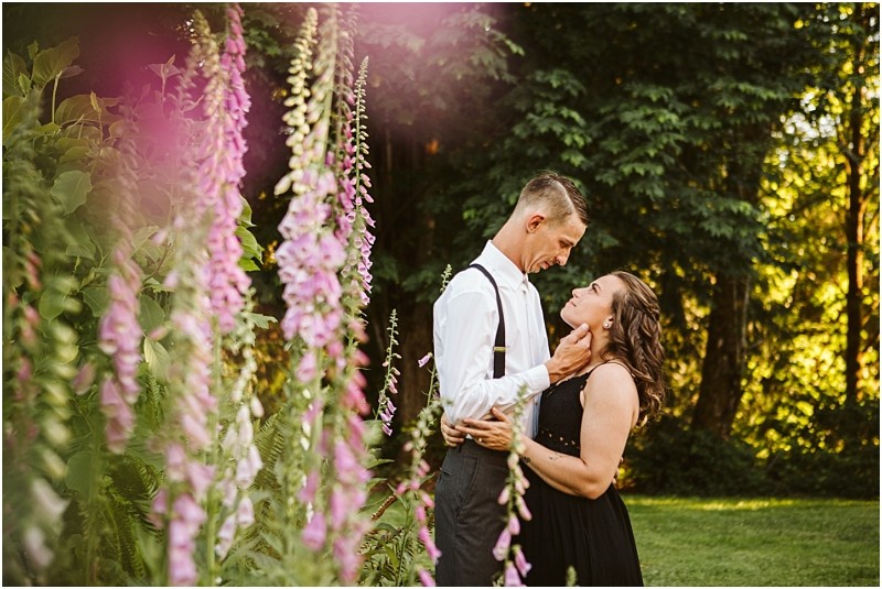 Premier Snohomish & Seattle Wedding Photographer | GSquared Weddings Photography A couple embraces lovingly in a sunlit garden, surrounded by tall pink flowers and lush greenery during their Snohomish Seattle wedding engagement. The man wears a white shirt and suspenders, while the woman gazes up at him affectionately in a black dress. 47.9129° N, 122.0982° W | Serving Snohomish, Seattle , Orlando & Beyond