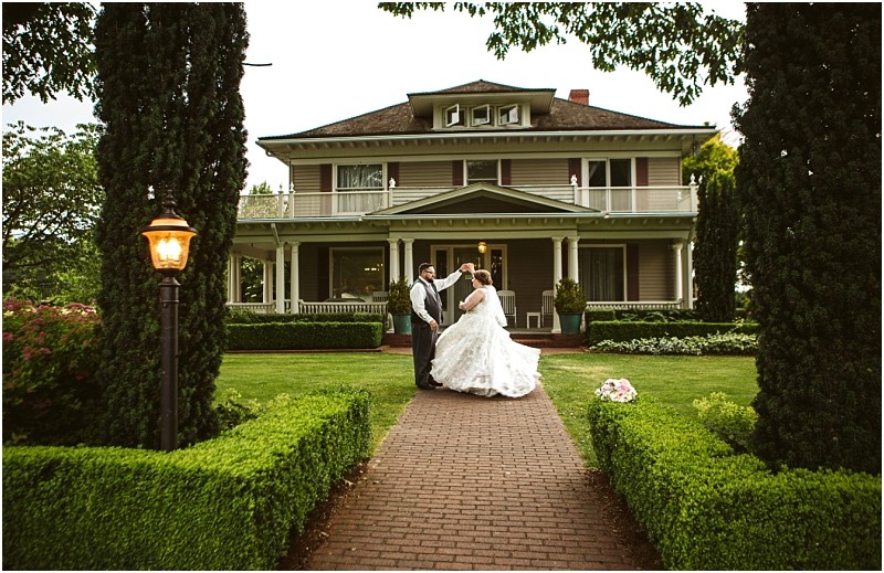 Premier Snohomish & Seattle Wedding Photographer | GSquared Weddings Photography A bride and groom dance on a brick pathway in front of a large, elegant house with a wraparound porch, surrounded by manicured gardens and tall trees—a romantic Snohomish wedding scene near Seattle. A lit lamp post glows on the left side of the path. 47.9129° N, 122.0982° W | Serving Snohomish, Seattle , Orlando & Beyond