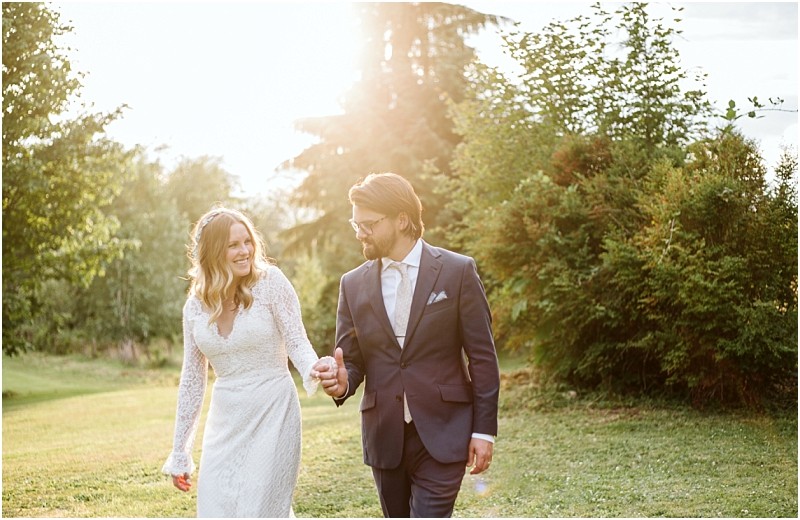 Premier Snohomish & Seattle Wedding Photographer | GSquared Weddings Photography A bride in a white lace dress and a groom in a navy suit walk hand in hand on a grassy lawn, smiling at each other, with sunlight streaming through the trees behind them during their Snohomish Seattle wedding. 47.9129° N, 122.0982° W | Serving Snohomish, Seattle , Orlando & Beyond