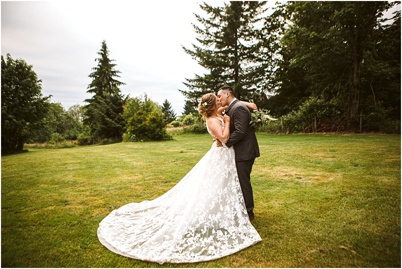 Premier Snohomish & Seattle Wedding Photographer | GSquared Weddings Photography A bride and groom embrace and kiss outdoors on a grassy lawn in Snohomish, surrounded by tall green trees. The bride wears a flowing white dress with a long train, while the groom is dressed in a dark suit—perfect for a Seattle wedding. 47.9129° N, 122.0982° W | Serving Snohomish, Seattle , Orlando & Beyond