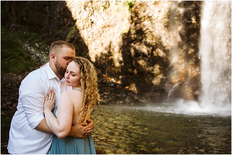 Premier Snohomish & Seattle Wedding Photographer | GSquared Weddings Photography A man embraces and kisses a woman on the forehead as they stand together by a waterfall near Snohomish, both appearing peaceful and affectionate. Sunlight highlights this romantic Seattle wedding engagement moment, with water cascading in the background. 47.9129° N, 122.0982° W | Serving Snohomish, Seattle , Orlando & Beyond