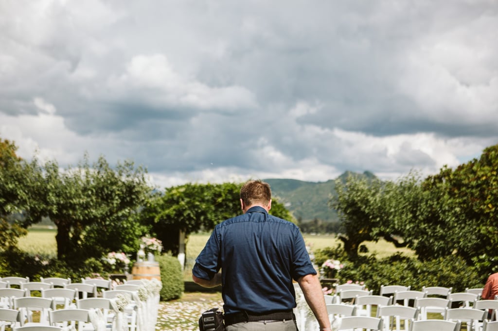Premier Snohomish & Seattle Wedding Photographer | GSquared Weddings Photography A man in a blue shirt walks between rows of empty white chairs set up outdoors for a Snohomish wedding, with green trees, mountains, and a cloudy sky in the background. 47.9129° N, 122.0982° W | Serving Snohomish, Seattle , Orlando & Beyond