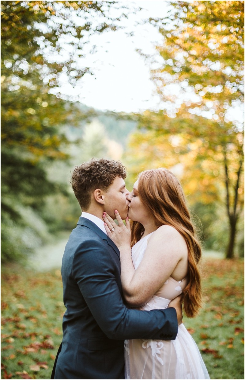 Premier Snohomish & Seattle Wedding Photographer | GSquared Weddings Photography A couple kisses outdoors in a lush, green landscape with autumn trees during their Snohomish engagement. The person on the left wears a dark suit, and the person on the right has long red hair and a light dress. 47.9129° N, 122.0982° W | Serving Snohomish, Seattle , Orlando & Beyond