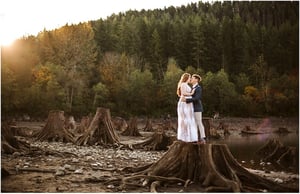 Premier Snohomish & Seattle Wedding Photographer | GSquared Weddings Photography A couple stands on a large tree stump in a rocky, dried lakebed near Snohomish, embracing at sunset with a forested hillside in the background—perfect for a Seattle wedding engagement session. 47.9129° N, 122.0982° W | Serving Snohomish, Seattle , Orlando & Beyond