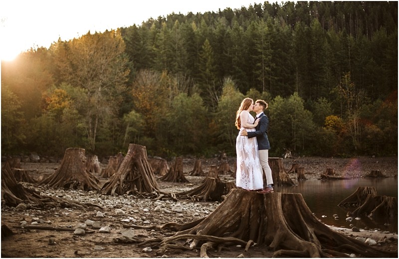 Premier Snohomish & Seattle Wedding Photographer | GSquared Weddings Photography A couple stands on a large tree stump in a rocky, dried lakebed near Snohomish, embracing at sunset with a forested hillside in the background—perfect for a Seattle wedding engagement session. 47.9129° N, 122.0982° W | Serving Snohomish, Seattle , Orlando & Beyond