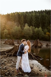 Premier Snohomish & Seattle Wedding Photographer | GSquared Weddings Photography A couple dressed in formal attire embraces and kisses on a rocky lakeshore near Snohomish, with tree stumps and a dense pine forest in the background. The soft light suggests either sunrise or sunset, capturing a romantic Seattle wedding engagement moment. 47.9129° N, 122.0982° W | Serving Snohomish, Seattle , Orlando & Beyond