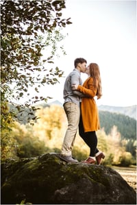 Premier Snohomish & Seattle Wedding Photographer | GSquared Weddings Photography A couple stands on a large mossy rock, embracing and kissing outdoors during their Snohomish engagement. The woman wears a long orange coat and the man wears a gray shirt and khaki pants, with trees and mountains in the background. 47.9129° N, 122.0982° W | Serving Snohomish, Seattle , Orlando & Beyond