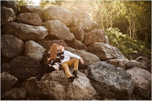 Premier Snohomish & Seattle Wedding Photographer | GSquared Weddings Photography A couple sits on large rocks outdoors in Snohomish, surrounded by greenery and sunlight. Sharing a kiss, the woman’s long red hair shines while the man, dressed in tan pants and a white shirt, creates a romantic Seattle engagement moment. 47.9129° N, 122.0982° W | Serving Snohomish, Seattle , Orlando & Beyond