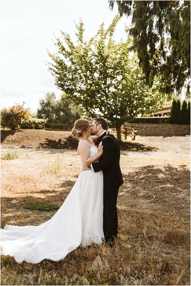 Premier Snohomish & Seattle Wedding Photographer | GSquared Weddings Photography A Snohomish bride and groom share a kiss outdoors on a sunny day. The bride wears a white strapless gown, and the groom is in a black suit. They stand in a grassy area with trees and a stone wall behind them at their Seattle wedding. 47.9129° N, 122.0982° W | Serving Snohomish, Seattle , Orlando & Beyond