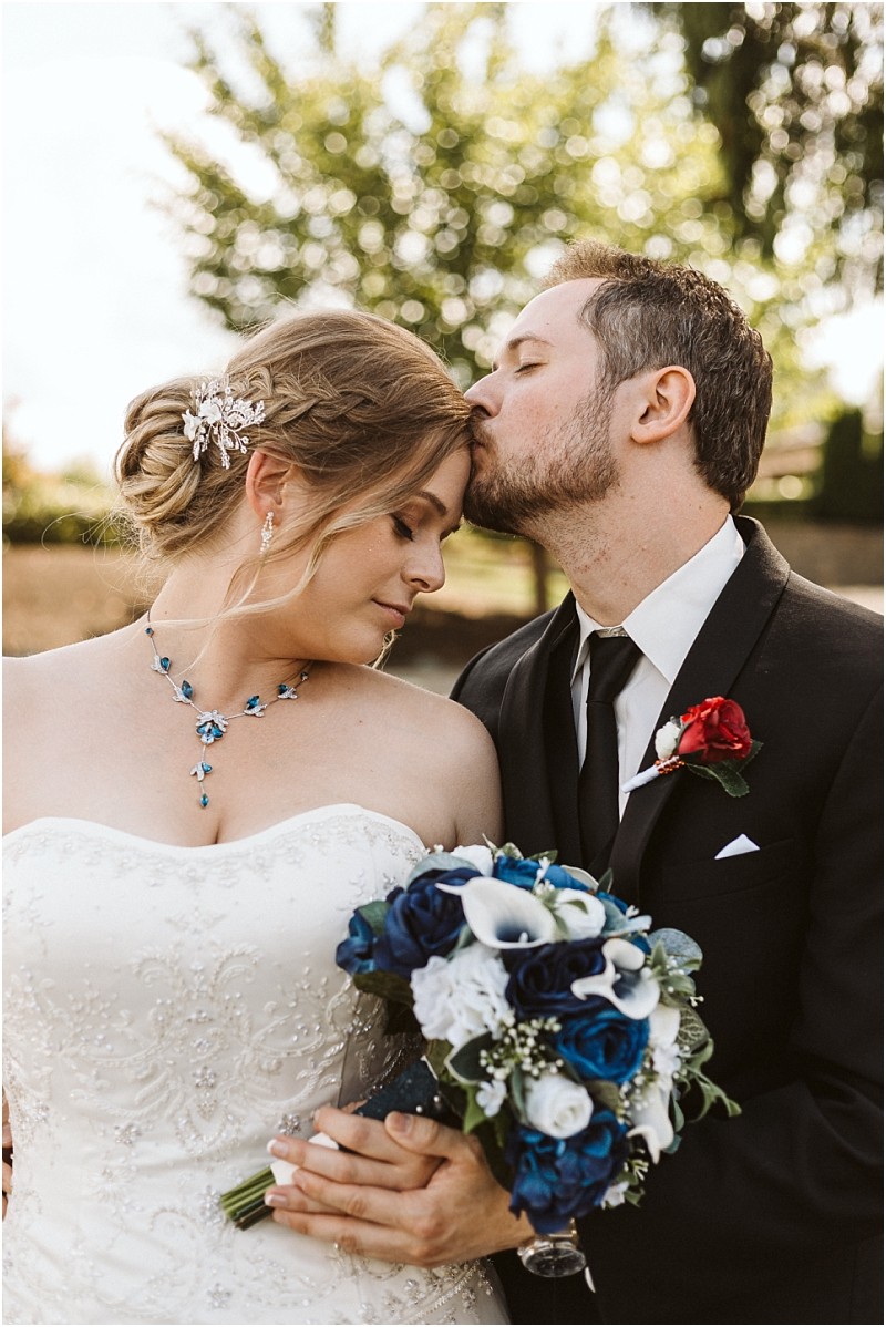Premier Snohomish & Seattle Wedding Photographer | GSquared Weddings Photography A groom in a black suit gently kisses the forehead of his bride in a strapless white dress holding blue and white flowers. This romantic moment is captured outdoors amid lush greenery at their Snohomish Seattle wedding engagement. 47.9129° N, 122.0982° W | Serving Snohomish, Seattle , Orlando & Beyond